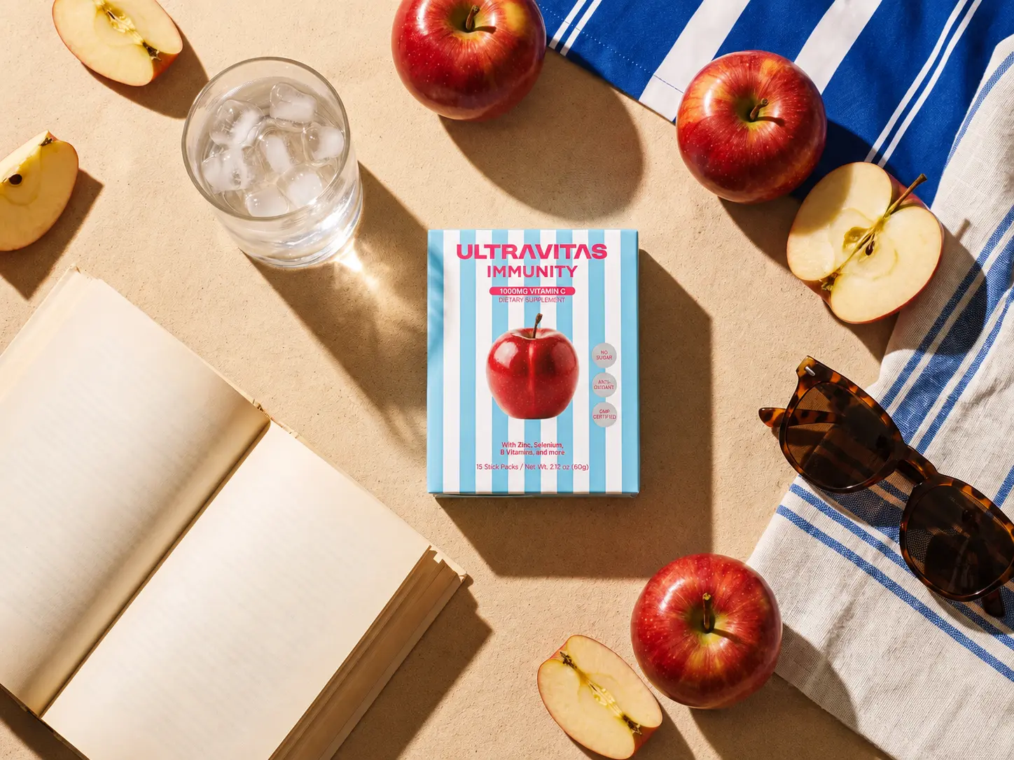 UltraVitas Immunity apple-flavored ultra-dose vitamin C powder sticks with apples, water, sunglasses, and a book.
