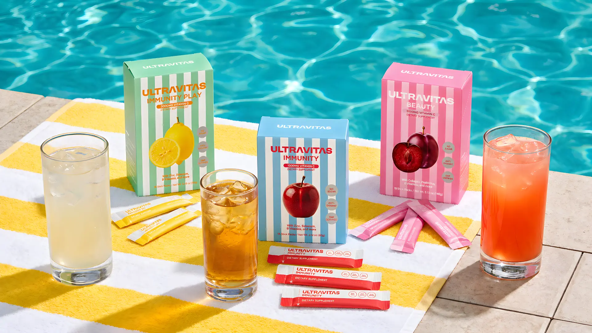 Three UltraVitas vitamin stick formulas displayed beside fruit-flavored drinks at a swimming pool.